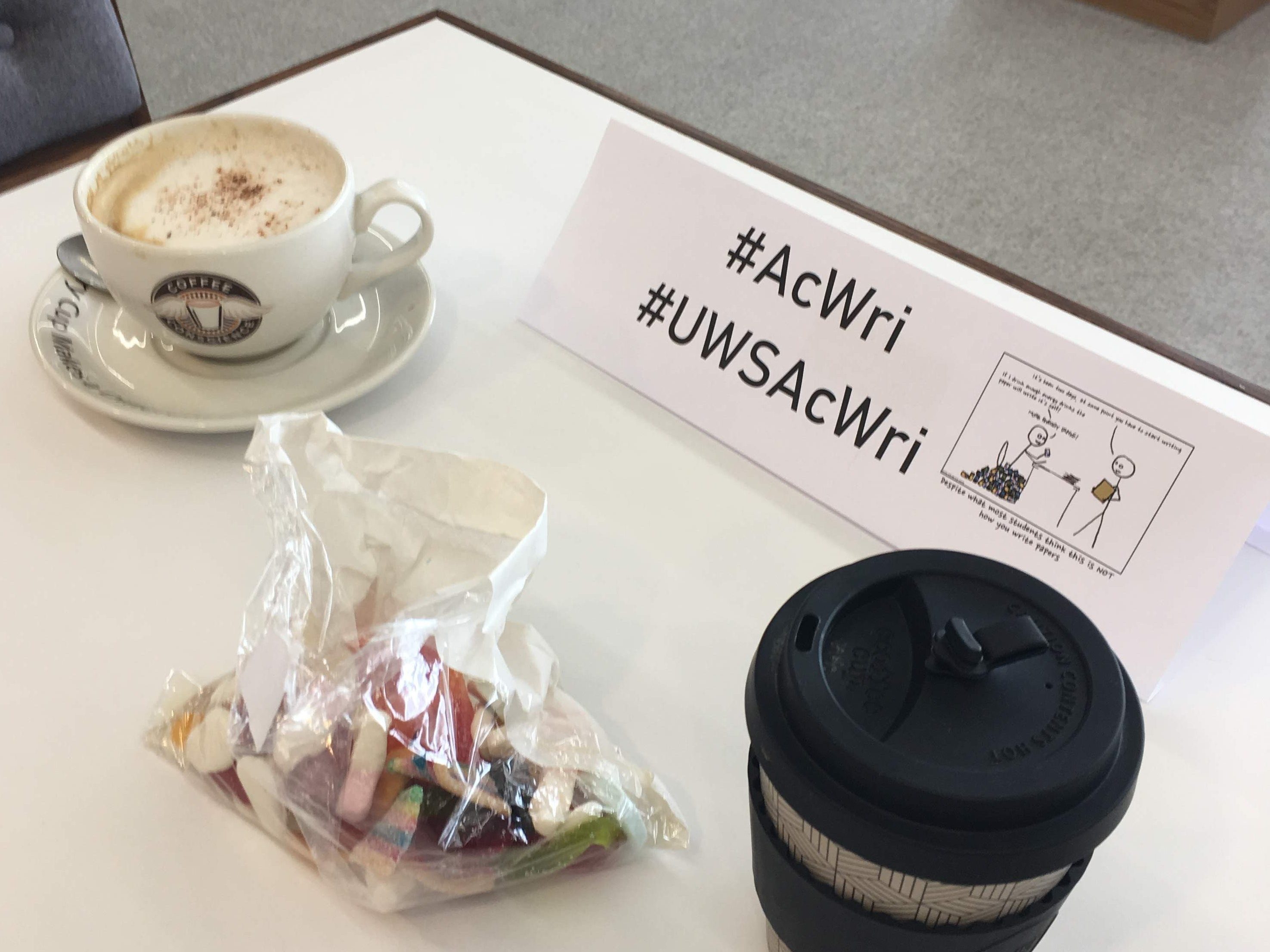 Two coffee cups on a table (one take away cup, one a ceramic one), a bag of sweets and the a sign advertising the writing group. 